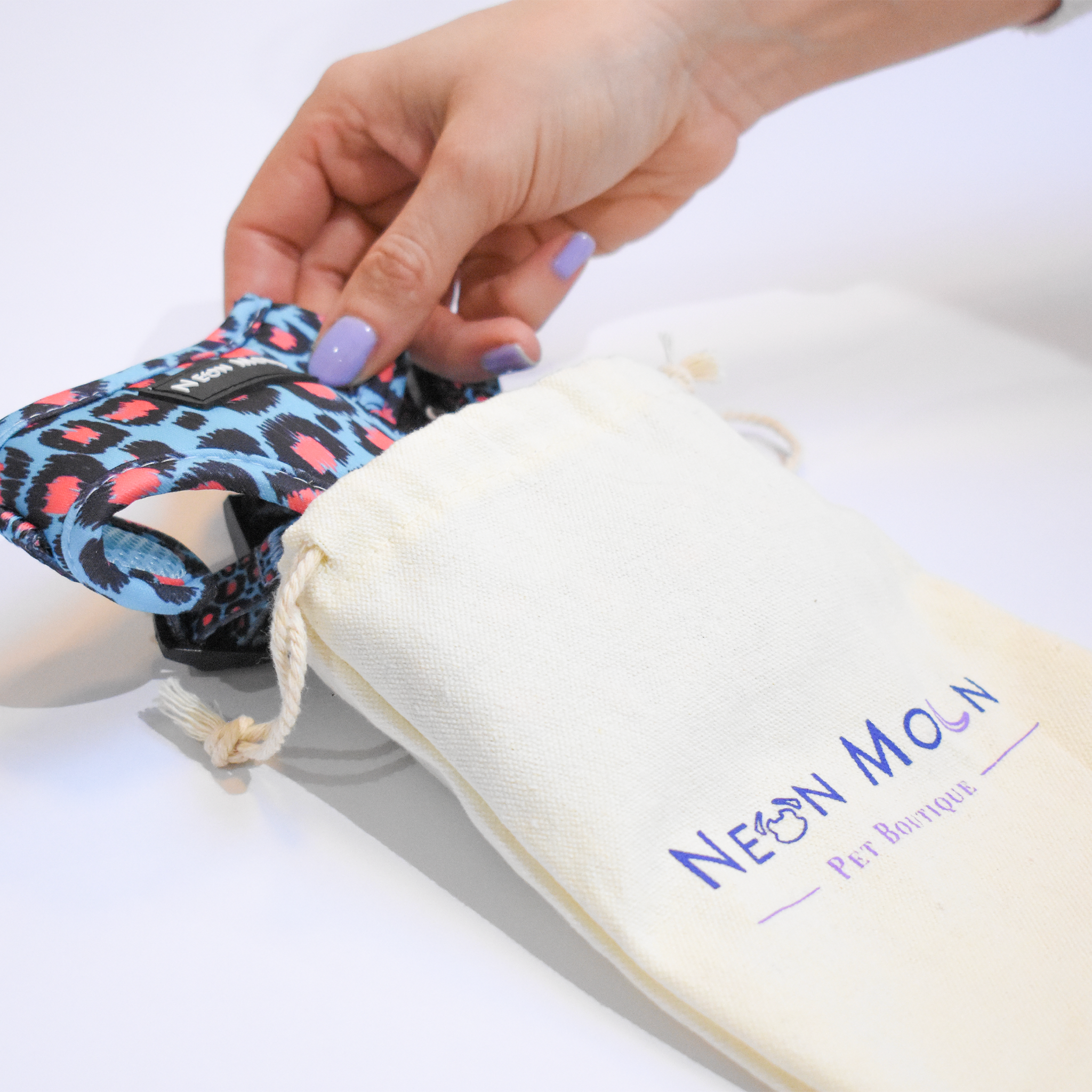 Hand holding a leopard print dog harness next to a white pouch with 'Neon Moon' branding on a light background.